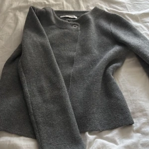 Grå kofta från Zara i ullmix - Mysig grå kofta från Zara i ullblandning. Enkel design med rund halsringning och en knapp framtill. Perfekt för lager-på-lager och passar till allt. Långärmad och i en klassisk, rak passform.