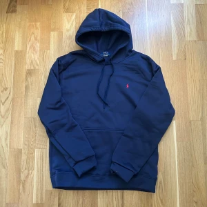 Marinblå hoodie från Polo Ralph Lauren - Marinblå hoodie från Polo Ralph Lauren med klassisk huva och snörning. Tröjan har en broderad röd logga på bröstet och en stor magficka. Tillverkad i mjuk bomullsmix och har långa ärmar med muddar. Perfekt för en chill och stilren look.