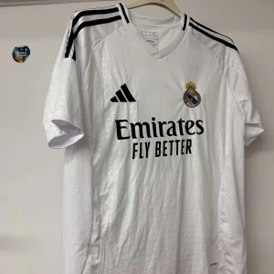 Real Madrid Mbappé #9 fotbollströja - Säljer en vit Real Madrid fotbollströja från Adidas med Mbappé och nummer 9 på ryggen. Tröjan har svarta detaljer, korta ärmar och klubbmärke på bröstet. Materialet är lätt och ventilerande polyester, perfekt för match eller träning.