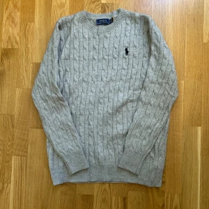 Grå kabelstickad tröja Polo Ralph Lauren - Kabelstickad grå tröja från Polo Ralph Lauren med klassisk rund halsringning och broderad svart logga på bröstet. Tröjan är långärmad och har ribbade muddar vid ärmslut och nederkant. Perfekt för lager-på-lager under kyliga dagar.