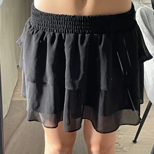 Svart volangkjol  - Säljer en svart kortkjol med volanger och bred resår i midjan. Kjolen är luftig och har ett tunt, lätt material som ger ett snyggt fall. Perfekt för dig som gillar en enkel men trendig look.