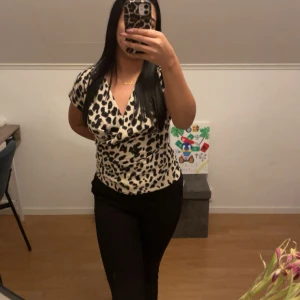 Leopardmönstrad omlottopp i beige och svart - Säljer en snygg omlottopp med leopardmönster i beige och svart. Toppen har korta ärmar, djup v-ringning och figurnära passform. Perfekt för dig som vill sticka ut med ett trendigt djurmönster. Materialet känns mjukt och följsamt mot kroppen.
