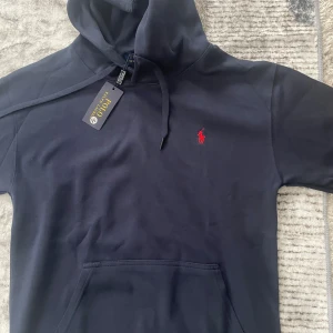 Mörkblå hoodie från Polo Ralph Lauren - Snygg mörkblå hoodie från Polo Ralph Lauren med klassisk röd logobrodyr på bröstet. Tröjan har huva med snörning, stor magficka och långa ärmar. Perfekt för dig som gillar stilrena och bekväma plagg med premiumkänsla.