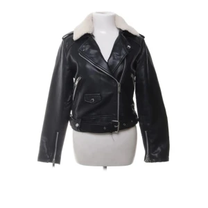 Svart skinnjacka med vit krage - Snygg svart skinnjacka med vit teddykrage och coola detaljer som dragkedjor, bälte i midjan och fickor med lock. Jackan har en klassisk bikerstil och är långärmad med en croppad passform. Perfekt för dig som vill ha en edgy look.