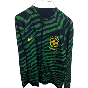 Nike Brasil långärmad fotbollströja M - Snygg långärmad fotbollströja från Nike för Brasilien, med marinblå och gröna zebraränder samt gul Nike-logga och Brasilien-emblem på bröstet. Slim fit-modell i lätt och sportigt material, perfekt för dig som vill sticka ut på planen.