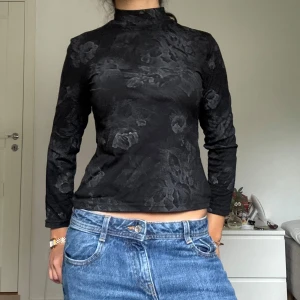 Svart blommig polotröja / festtop - Säljer en svart polotröja med diskret blommigt mönster. Tröjan har lång ärm och en tight passform med hög hals.