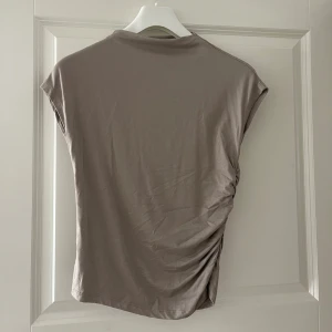 Beige ärmlös topp med rynkdetalj - Säljer en beige ärmlös topp i mjukt material med snygg rynkdetalj på ena sidan. Toppen har en enkel och stilren design med rund hals och loose passform, perfekt för en avslappnad look.