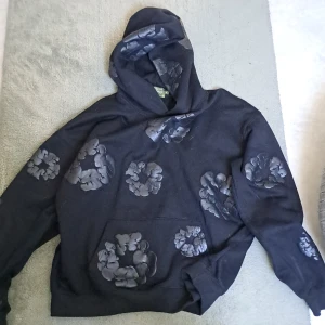 Svart hoodie från Denim Tears med blommor - Svart hoodie från Denim Tears med unika blommiga mönster i mörkare nyans över hela tröjan. Tröjan har huva, känguruficka och är långärmad. Perfekt för dig som vill sticka ut med en cool och kreativ streetwear-look.
