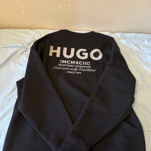 Svart Sweatshirt från Hugo - Svart sweatshirt från Hugo med vit logga på bröstet och stort tryck på ryggen. Tröjan har långa ärmar och är gjord i mjuk bomull. Perfekt för dig som gillar märkeskläder. *Endast använd en gång*
