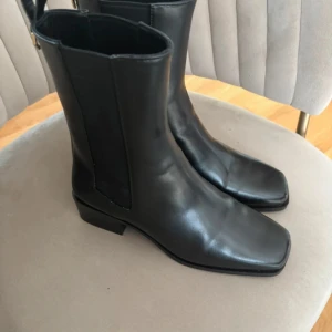 Svarta chelsea boots med fyrkantig tå - Snygga svarta chelsea boots i skinn med fyrkantig tå och låg klack. Bootsens minimalistiska design har elastiska paneler på sidorna och en guldfärgad detalj baktill. Perfekta för dig som gillar stilrena och trendiga skor.