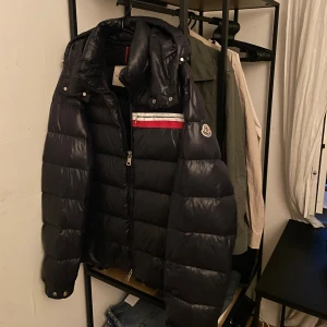 Moncler mounier dunjacka med huva - Säljer en mörkblå dunjacka från Moncler med avtagbar huva och snygga detaljer i rött och vitt på bröstet. Jackan har Moncler-logga på ärmen, dragkedja framtill och quiltad design som ger en schysst puffer-look. Perfekt för kalla dagar!