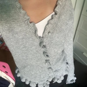 Grå kofta med volang och pärlknappar - Superfin grå kofta med volangkant längs halsringning, nederkant och ärmslut. Stängs med pärlknappar framtill och är stickad i mjukt material. Perfekt för dig som gillar söta detaljer och vill ha en mysig, men ändå stylish look.