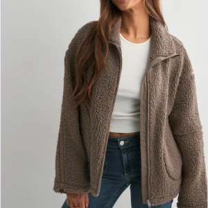 Brun pile jacka från Nelly XS - Mysig brun teddyjacka från Nelly i storlek XS. Jackan har en oversized look, dragkedja framtill och stora fickor. Perfekt för kyliga dagar med sitt fluffiga material och höga krage. Jackan är helt ny och har inte använts☺️