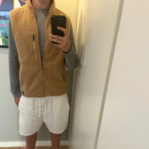 Beige fleeceväst med dragkedja - Säljer en beige fleeceväst  med hög krage och tre fickor med dragkedja. Västen har en mjuk och mysig känsla, perfekt att slänga på sig över en hoodie. Enkel och stilren design med diskret logga nertill.