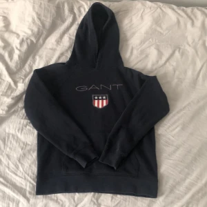 Mörkblå hoodie från GANT - Säljer en Mörkblå hoodie från GANT med broderad logga på bröstet. Väldigt basic för en både snygg och avslappnad stil. Kan bäras på sena sommar kvällar och vinter. Sjukt mjuk och skön! Priset är inte hugget i sten! Tveka aldrig med att höra av dig.