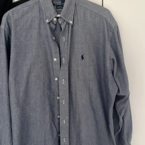 Blå skjorta från Polo Ralph Lauren - Snygg blå skjorta från Polo Ralph Lauren med klassisk button-down krage och broderad logga på bröstet. Skjortan har lång ärm, knappar framtill och är tillverkad i mjuk bomull. Perfekt för en clean och stilren look. Är väldigt stor i storlek är som XL säljer för 250