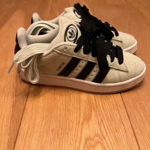 Adidas Campus sneakers i mocka - Snygga Adidas Campus sneakers i ljusbeige mocka med svarta ränder och svart Adidas-logga på plösen. Skorna har både svarta och vita skosnören så du kan variera stilen. Klassisk rund tå och platt sula, perfekt för en streetwear-look. Helt nya bara testade 