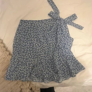 Blå blommig kjol med volang - Supersöt blå kortkjol med vitt blommönster och volangkant. Kjolen har dragkedja på sidan och även en rosett på sidan. 