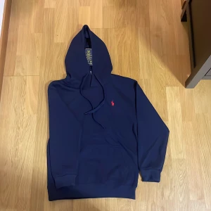  Polo Ralph Lauren hoodie - Säljer en marinblå hoodie från Polo Ralph Lauren med klassisk röd broderad logga på bröstet. Tröjan har huva med snörning, känguruficka och ribbade muddar. Perfekt för dig som gillar stilrena och bekväma plagg.