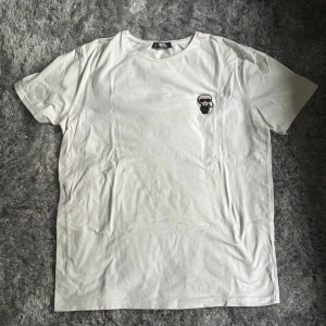 Vit t-shirt från Karl Lagerfeld - Vit t-shirt från Karl Lagerfeld med klassisk passform och rund hals. Skulle säga passar L också.  På bröstet finns en liten broderad patch med Karl Lagerfeld-figur i svart och vitt. Perfekt basic med en cool detalj som sticker ut.
