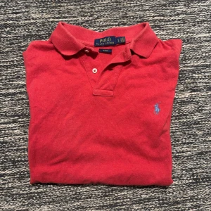 Röd pikétröja Polo Ralph Lauren - Röd pikétröja från Polo Ralph Lauren i slim fit-modell. Klassisk krage, knäppning framtill och broderad blå logga på bröstet. Tillverkad i bomull och passar perfekt för en clean och sportig stil.