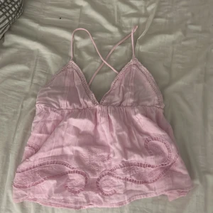 Ljusrosa linnetopp från Gina Tricot - Supersöt ljusrosa linnetopp från Gina Tricot med tunna axelband som korsas i ryggen. Toppen har broderade detaljer och spets längs v-ringningen, samt en flowy passform. Den är helt ny och oanvänd.