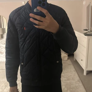 Blå jacka Polo Ralph Lauren - Quiltad blå jacka från Polo Ralph Lauren med röd insida och klassisk logga på bröstet. Jackan har dragkedja framtill, ribbade muddar och diskreta knappar vid fickorna. Perfekt för höst och vinter, stilren och bekväm med snygg passform. Storlek L i barn (14-16)