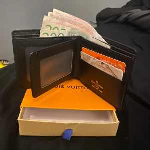 Lyxig svart rutig plånbok - Snygg svart plånbok i klassiskt rutigt mönster. Tillverkad i exklusivt skinn med flera kortfack, sedelfack och ett fönster för ID eller körkort. Kommer med box och har en stilren, modern look. Pengar och kort som syns på bild ingår inte