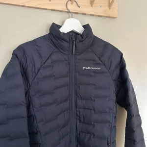 Mörkblå pufferjacka Peak Performance - Säljer en mörkblå pufferjacka från Peak Performance med hög krage och två dragkedjeförsedda fickor. Jackan har diskret logga på bröstet och ärmarna, och är tillverkad i ett syntetmaterial som håller värmen. Perfekt för kyliga dagar.
