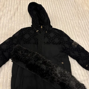 Svart pufferjacka Glorious Gangsta - Säljer en svart pufferjacka från Glorious Gangsta med snyggt mönster på axlar och ärmar. Jackan har huva med avtagbar fuskpäls, gulddetaljer och dragkedja framtill. Perfekt för kalla dagar och riktigt stilren look.