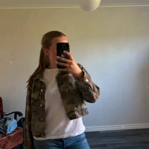 Croppad camo jeansjacka - Säljer en croppad jeansjacka med camouflagemönster i gröna och bruna toner. Jackan har långa ärmar, knappar framtill och klassiska fickor på bröstet. Perfekt för dig som vill ha en edgy och cool stil.