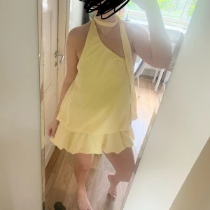 Gul kortklänning med volanger - Superfin ljusgul kortklänning i luftig modell med volanger och halterneck. Klänningen har öppen rygg och en snygg scarf-detalj runt halsen. Perfekt för sommardagar när du vill sticka ut lite extra.