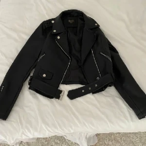 Svart skinnjacka med bälte - Svart skinnjacka med klassisk bikerstil, silvriga dragkedjor och knappar. Jackan har bälte i midjan och croppad passform. Perfekt för dig som vill ha en edgy look med snygga detaljer.