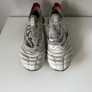 Mizuno Morelia Neo fotbollsskor vita - Säljer ett par Mizuno Morelia Neo Made in Japan  fotbollsskor i vitt skinn med svarta detaljer och röd häl. Skorna har snörning, rund tå och klassiska dobbar. Ovandelen är i mjukt läder och mesh för extra komfort och känsla på planen. Ett par riktigt sköna och lätta läderskor som är sjukt snygga. Lite defekter förekommer på tån längst fram men fungerar alldelses utmärkt använda fåtal gånger men den gångerna va på en konstgräs plan med svarta pluppar som gjorde skorna lite smutsiga.