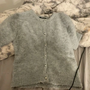 Ljusgrå kofta från Gina Tricot XS - Supermjuk ljusgrå kofta från Gina Tricot i fluffigt stickat material. Koftan har korta ärmar och pärlknappar framtill som ger en söt touch. Perfekt att slänga över en topp för extra mysfaktor.