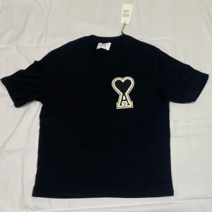 Svart t-shirt från Ami Paris med hjärtdetalj - Svart t-shirt från Ami Paris med klassisk passform och korta ärmar. På bröstet sitter en stor broderad logga i form av ett hjärta och bokstaven A i vitt. Tillverkad i mjuk bomull som känns skön mot huden. Perfekt för en clean och stilren look.