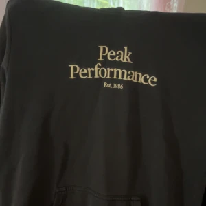 Mörkblå hoodie från Peak Performance - Mörkblå hoodie från Peak Performance med broderad logga och text på bröstet. Luva bak, klassisk känguruficka framtill och mjukt material som känns skönt mot huden. 