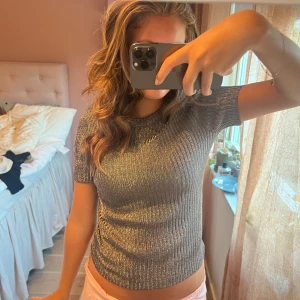 Glittrig ribbad silver topp - Säljer en glittrig ribbad topp i silver med korta ärmar och rund hals. Toppen har en tight passform och är tillverkad i ett stretchigt material som skimrar snyggt i ljuset. Perfekt för dig som vill sticka ut lite extra.