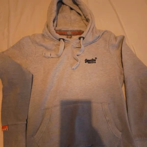 Grå hoodie från Superdry med huva - Säljer en ljusgrå/ljusblå hoodie från Superdry med broderad logga på bröstet. Tröjan har huva med snörning, känguruficka och ribbade muddar. Perfekt för dig som gillar en avslappnad och sportig stil. Pris kan diskuteras