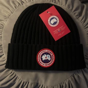 Svart ribbstickad mössa Canada Goose - Svart ribbstickad mössa från Canada Goose med klassisk uppvikt kant och broderad logotyp framtill. Tillverkad i mjukt ullmaterial som håller dig varm under kalla dagar. Perfekt accessoar för vintern med stilren och tidlös design.