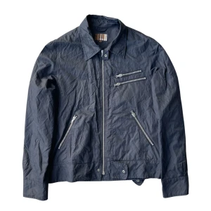 Svart vindjacka från Emillio  - Svart vindjacka från Emillio med coola silverfärgade dragkedjor på bröstet och vid fickorna. Jackan har klassisk krage, raka ärmar och en lätt, lite skrynklig look. Perfekt för dig som gillar stilrena detaljer och vill ha en jacka som sticker ut.