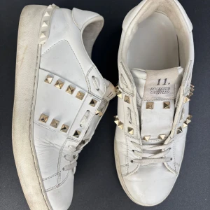 Valentino Rockstud vita sneakers - Snygga vita Valentino Rockstud sneakers i skinn med ikoniska guldiga nitar längs sidorna och hälen. Klassisk rund tå, snörning och platt sula. Perfekta för dig som vill sticka ut med en lyxig och edgy vibe.