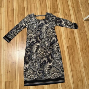 Mönstrad klänning från Happy Holly - Snygg långärmad klänning från Happy Holly i storlek 32/34. Klänningen har ett paisleyinspirerat mönster i svart, grått och beige med v-ringning och dekorativ detalj i nacken. Perfekt för dig som gillar unika prints och bekväm passform.