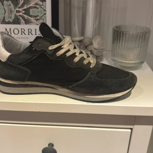 Philippe Model svarta sneakers  - Svarta sneakers från Philippe Model med detaljer i mocka och textil, vit hälkappa och vita snören. Sidan har en gulmarkerad sköldlogga. Snygg, sportig design med platt sula och rund tå. Perfekta för dig som gillar streetstyle och franska märken.