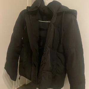 Svart pufferjacka från DeFacto - Svart pufferjacka från DeFacto med huva och dragsko. Jackan har en hög krage, dragkedja framtill och två stora fickor. Perfekt för kalla dagar när du vill hålla dig varm och ändå se snygg ut.