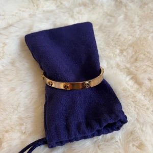 Guldigt armband från Tory Burch - Snyggt armband i guldton från Tory Burch med klassiska loggor runt om. Armbandet är stelt och har en stilren, polerad finish som ger en lyxig känsla. Perfekt accessoar för att lyfta din outfit med en touch av elegans.