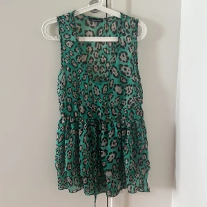 Grön leopardmönstrad kortklänning - Snygg grön kortklänning från Line Standard med leopardmönster. Klänningen är ärmlös, har v-ringning och ett tunt, luftigt material. Midjan markeras med knytband och kjolen har volang för extra flow. Passar som strand coverup 