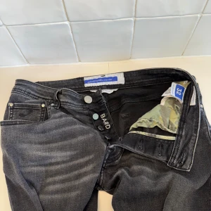 Jacob cohen jeans  - Snygga svarta jeans från Jacob Cohën i modell Bard. Jeansen har en klassisk femficksdesign, knäppning med knappar. Materialet är mjukt och stretchigt jeans, perfekt för dig som gillar stilrena och bekväma byxor. Loggan där bak har släppt lite! Bara att höra av sig till oss vid minsta fundering!! 