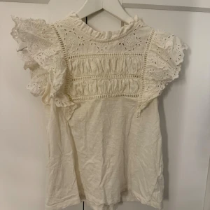 Vit blus med spets och volang - Säljer en söt vit blus med broderade spetsdetaljer och volang på axlarna. Blusen har en hög krage och är ärmlös med romantiska detaljer och hålmönster upptill. Perfekt för dig som gillar en feminin och trendig stil.