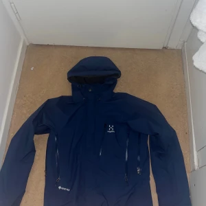  Haglöfs gortex jacka - Säljer en mörkblå vindjacka från Haglöfs med huva och flera praktiska dragkedjor framtill. Jackan är tillverkad i slitstark Gore-Tex och har långärmad design, perfekt för utomhusaktiviteter. Snygg och funktionell för dig som gillar friluftsliv. Passar S/M då man kan spänna in den till sin egna preferens.
